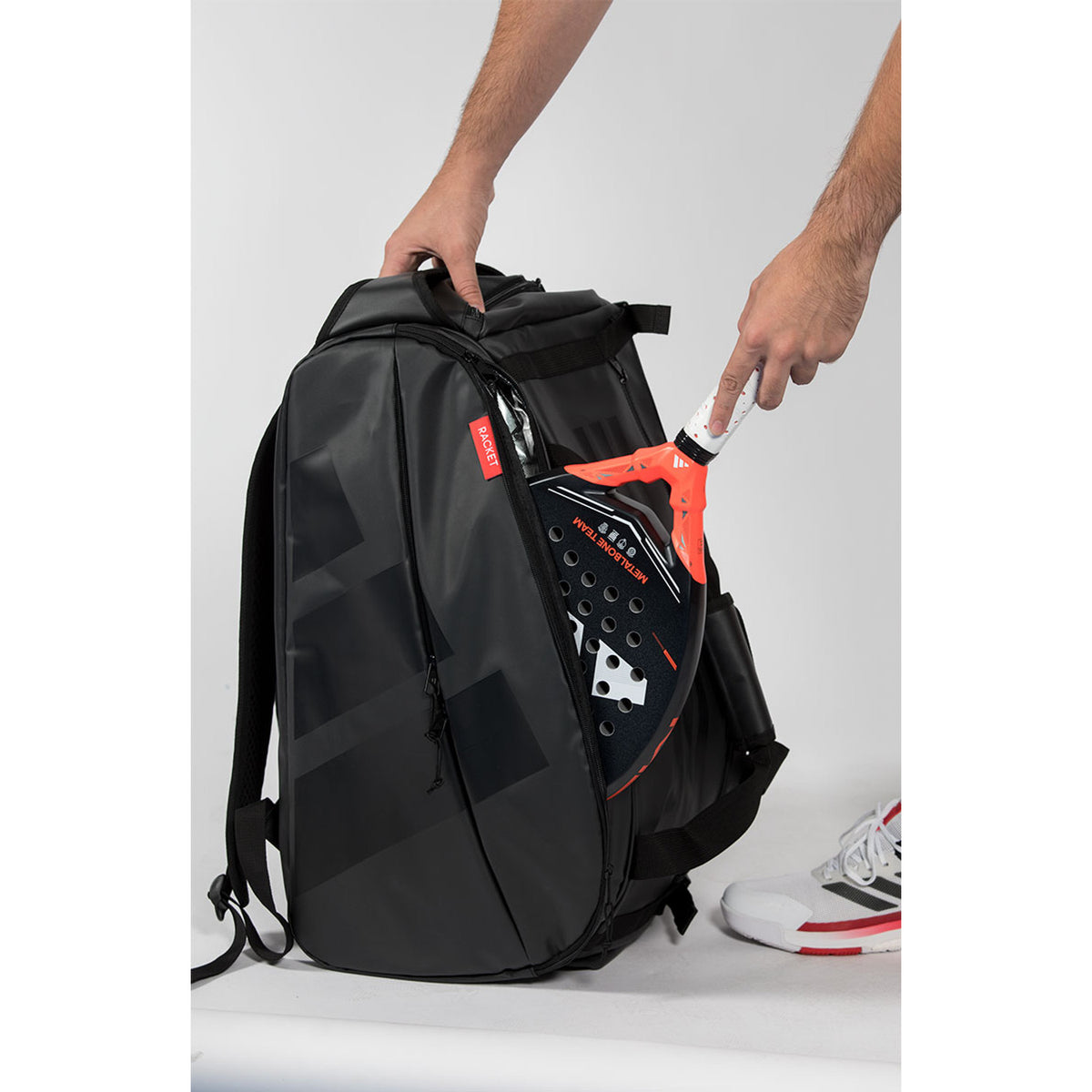 Hand opent het racketvak van de zwarte Adidas Multigame 2026 met oranjerode trekkers.