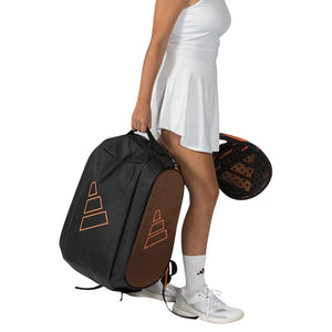 Speelster draagt de donkere Adidas Tour 2026 racketbag aan de hand naast haar racket.