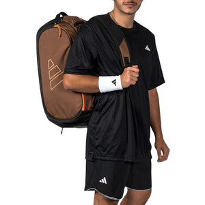 Man draagt de bruine Adidas Racket Bag Tour 2026 over zijn schouder in zwarte sportkleding.