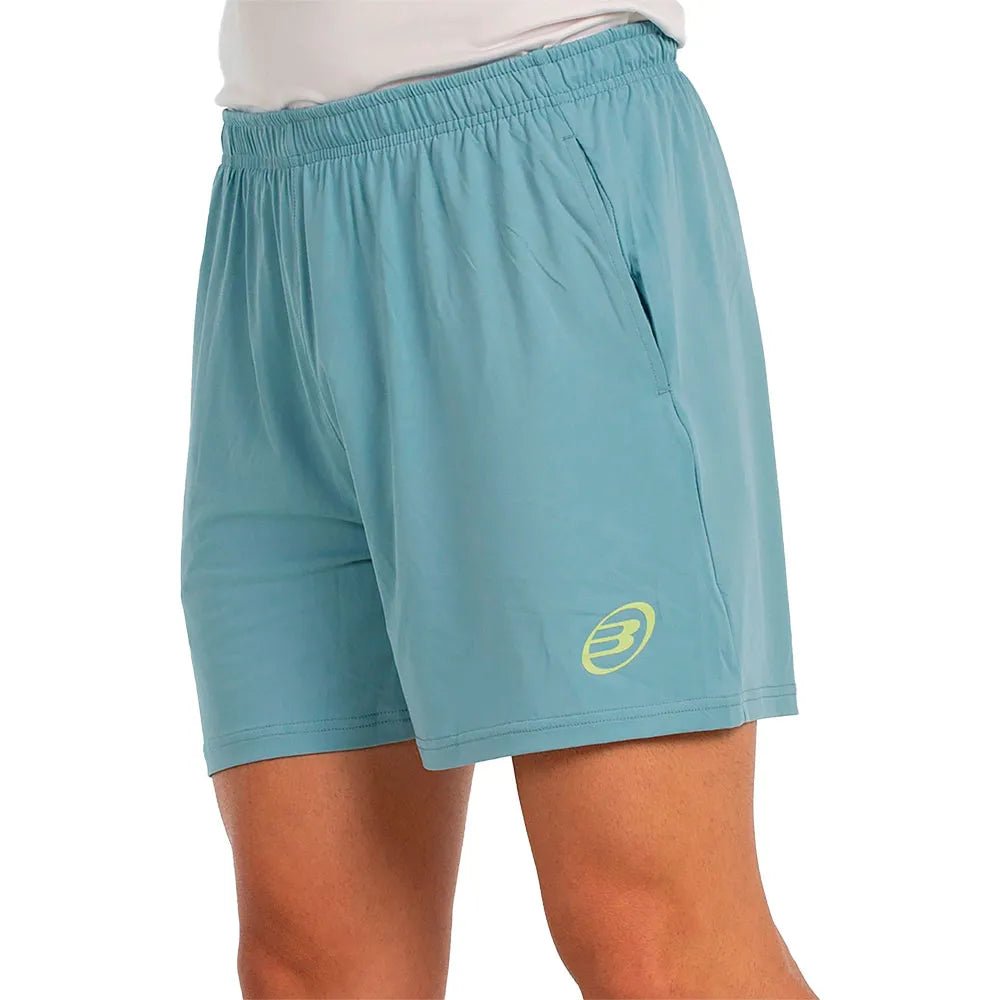 Model draagt de aquablauwe Bullpadel Short Lijar, schuin aanzicht met geel logo op de pijp